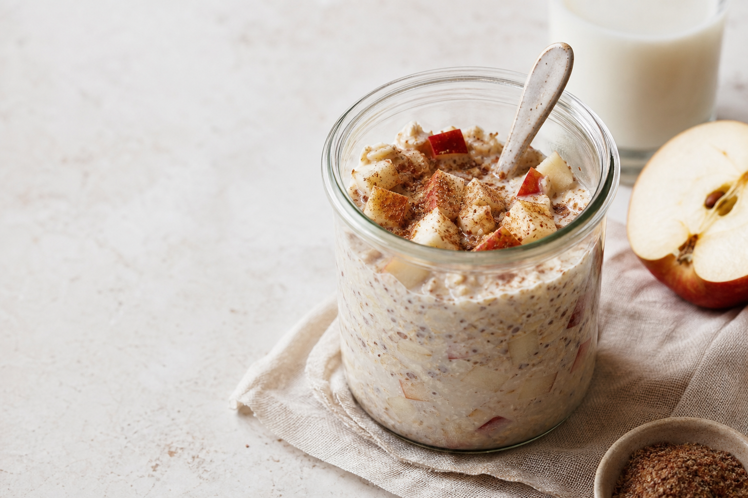 Glass jar of overnight oats with diced apple, ground flaxseed, and cinnamon beside soy milk